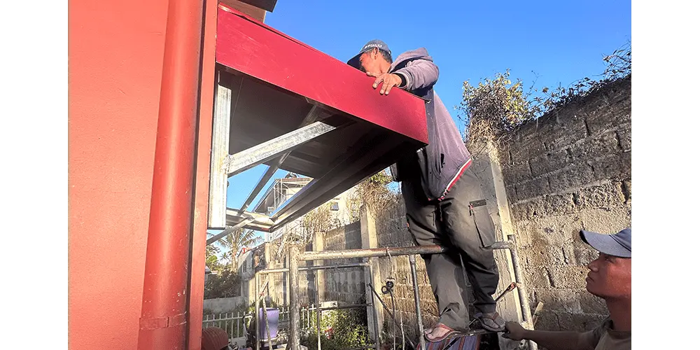 脚立に乗ってトタンを取り付けるフォアマン。青空が清々しい